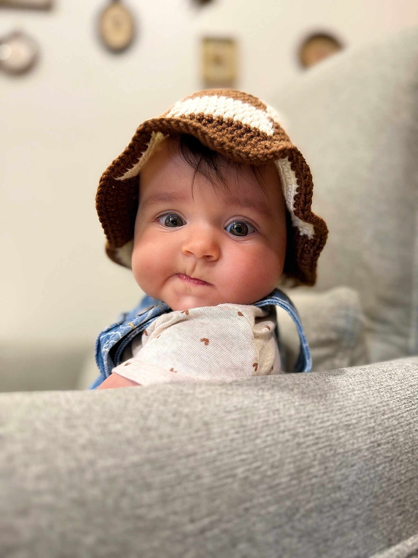 Olive is registered to the contest to win money with this photo: baby, child, face, hat, knitted_hat, brown, white, clothing, couch, furniture, indoor, portrait, cute, chubby_cheeks, looking_at_camera, soft_light, blurred_background, wall_decor, cozy, comfortable