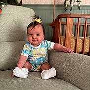 Olive is registered to the contest to win money with this photo: baby, child, infant, onesie, socks, couch, furniture, crib, mobile, indoor, cute, smile, hair_bow, seated, nursery, cozy, chubby_cheeks, person, home, soft_lighting
