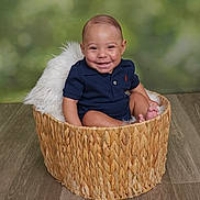 Sebastian is registered to the contest to win money with this photo: baby, smiling, basket, blue_clothing, indoor, flooring, fluffy_blanket, child, cute, portrait, happy, sitting, woven_basket, infant, studio_photo, green_background, casual_clothing, feet, one_person, young_child