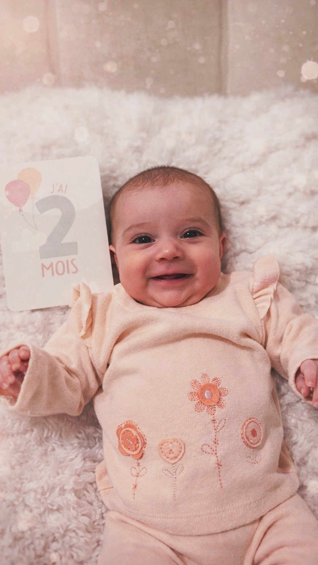 Joy a rejoint le concours — aidez-le/la à gagner de superbes lots ! baby, infant, smile, face, blanket, clothing, soft_toys, milestone_card, cute, portrait, indoors, child, baby_girl, floral_pattern, happy, newborn, lying_down, warm_colors, cozy, closeup