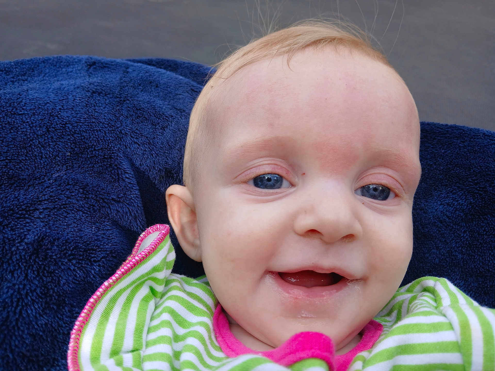 Olivia is registered to the contest to win money with this photo: baby, infant, close_up, drooling, smiling, blue_eyes, navy_towel, blanket, striped_clothing, green_and_white, face, ear, forehead, portrait, candid, outdoor, clothing, happy, mouth, eye_contact