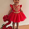 barefoot, beige_carpet, birthday, child, cute, flower_decor, girl, gold_embroidery, happy, headband, holding_prop, number_two, plain_wall, portrait, red_dress, roses, ruffles, smile, standing, toddler