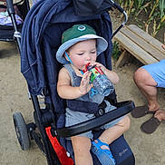 Sasha a rejoint le concours — aidez-le/la à gagner de superbes lots ! toddler, child, stroller, hat, water_bottle, outdoor, bench, dirt_path, sandals, clothing, person, baby, sun_hat, footwear, greenery, adult, legs, relaxing, summer, daylight