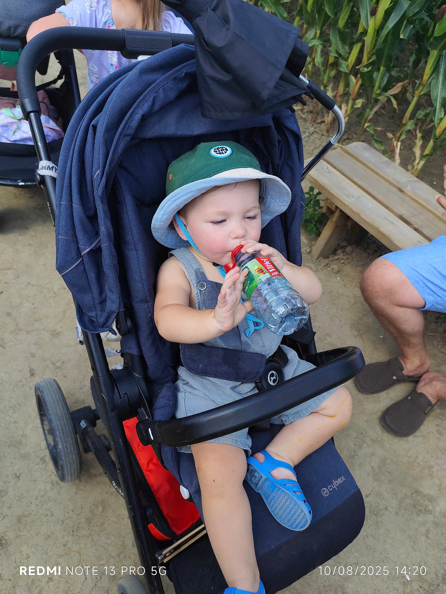 Sasha a rejoint le concours — aidez-le/la à gagner de superbes lots ! toddler, child, stroller, hat, water_bottle, outdoor, bench, dirt_path, sandals, clothing, person, baby, sun_hat, footwear, greenery, adult, legs, relaxing, summer, daylight