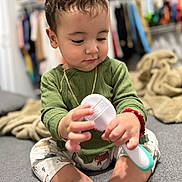 Liam Matías is registered to the contest to win money with this photo: toddler, child, baby, green_shirt, patterned_pants, sitting, handheld_object, indoor, soft_focus, blanket, clothing, barefoot, bracelet, necklace, floor, toys, cute, playing, person, portrait