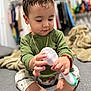 toddler, child, baby, green_shirt, patterned_pants, sitting, handheld_object, indoor, soft_focus, blanket, clothing, barefoot, bracelet, necklace, floor, toys, cute, playing, person, portrait