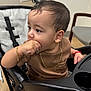toddler, child, stroller, indoor, brown_clothing, gold_chain, gold_bracelet, hand_in_mouth, curly_hair, face, chair, window, floor, expression, person, portrait, seated, contemplative, baby, modern