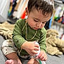 toddler, child, green_shirt, barefoot, carpet, indoor, toy, focused, pants, patterned_pants, blanket, floor, hair, gold_chain, hand, feet, room, closet, baby, sitting