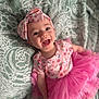 baby, child, smiling, headband, pink_tutu, floral_pattern, happy, infant, clothing, blanket, portrait, cute, joyful, face, person, toddler, apparel, fabric, lying_down, indoor