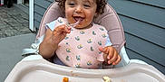 Viviana is registered to the contest to win money with this photo: toddler, child, high_chair, bib, pasta, messy, food, eating, happy, curly_hair, outdoor, patio, gray_siding, plastic_tray, fork, smiling, clothing, young_child, meal_time, baby
