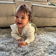 Viviana is registered to the contest to win money with this photo: baby, child, crawling, smile, happy, indoor, rug, carpet, furniture, sofa, hair_clip, curly_hair, white_shirt, green_pants, cute, portrait, person, home, floor, bright