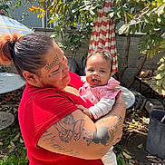 Miracle joined the competition — help win amazing prizes! baby, adult, tattoos, red_shirt, outdoor, garden, umbrella, greenery, smiling, holding, cute, person, infant, happy, daylight, nature, casual, portrait, family, love