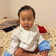 Anand a rejoint le concours — aidez-le/la à gagner de superbes lots ! baby, child, infant, sitting, bed, blanket, white_clothing, blue_cuffs, face, eyes, hair_clip, chubby_cheeks, toes, legs, indoor, wall, pattern_bedspread, looking_at_camera, portrait, cute