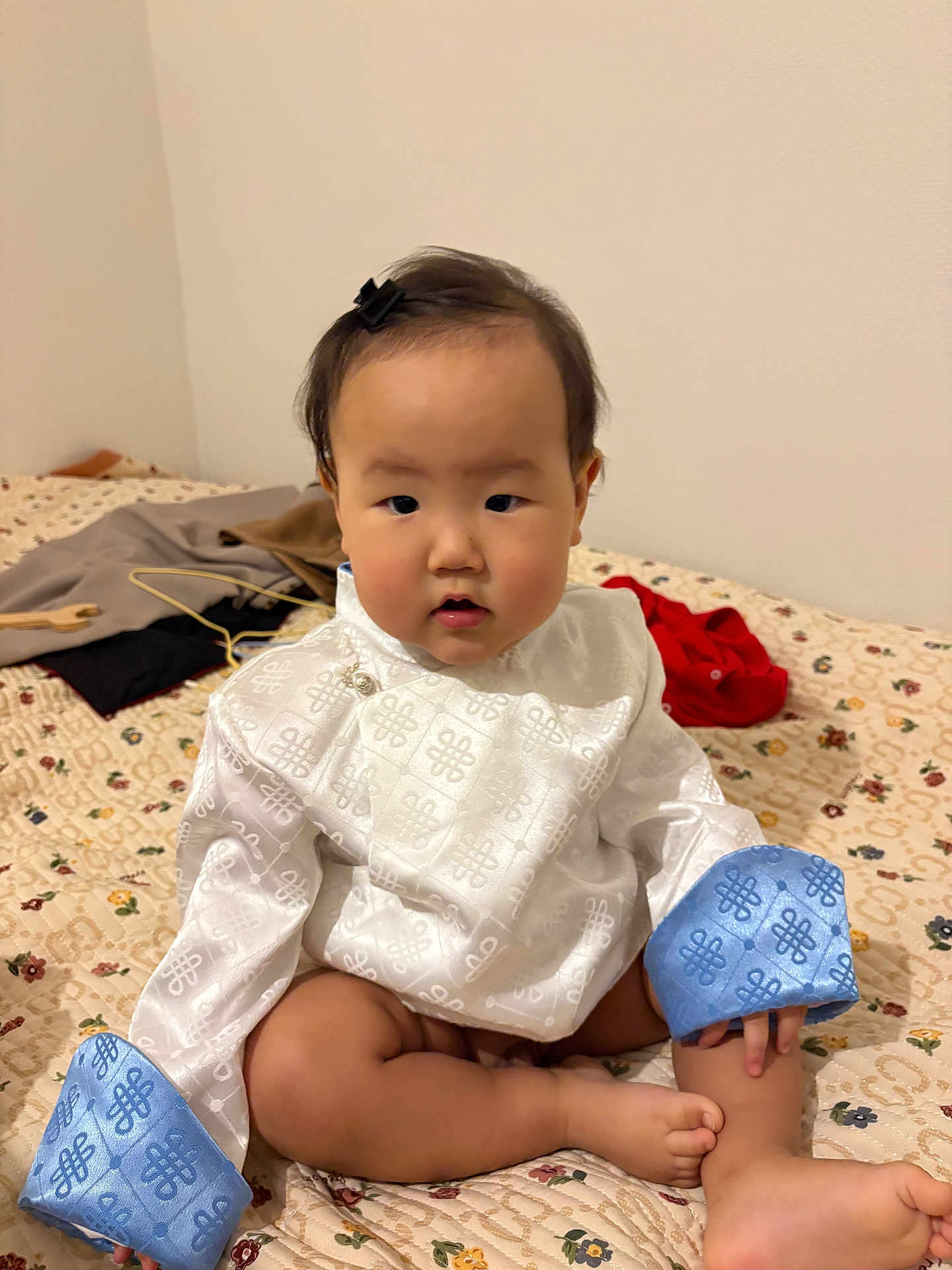 Anand a rejoint le concours — aidez-le/la à gagner de superbes lots ! baby, child, infant, sitting, bed, blanket, white_clothing, blue_cuffs, face, eyes, hair_clip, chubby_cheeks, toes, legs, indoor, wall, pattern_bedspread, looking_at_camera, portrait, cute