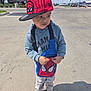 child, toddler, boy, spiderman_hat, cap, hoodie, sling_bag, bag, street, pavement, shoes, sneakers, standing, outdoor, sunny, shadow, urban, looking_right, serious_expression, portrait