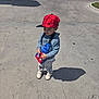 child, toddler, red_hat, cap, hoodie, crossbody_bag, superhero_bag, sneakers, standing, shadow, pavement, sidewalk, parking_lot, outdoor, sunlight, casual_clothing, portrait, candid, urban, smile