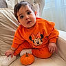 baby, child, infant, portrait, sitting, orange_outfit, minnie_mouse, pumpkin, sneaker, couch, cushions, window, indoor, daylight, wide_eyed, surprised, jewelry, bracelet, cross_necklace, autumn