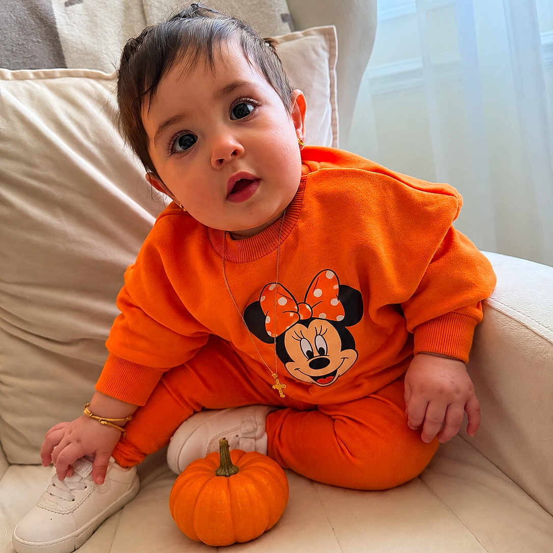 Amelia joined the competition — help win amazing prizes! baby, child, infant, portrait, sitting, orange_outfit, minnie_mouse, pumpkin, sneaker, couch, cushions, window, indoor, daylight, wide_eyed, surprised, jewelry, bracelet, cross_necklace, autumn