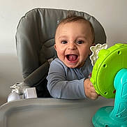 Khalil joined the competition — help win amazing prizes! baby, child, high_chair, toy, smile, happy, indoor, infant, person, playful, blue_clothing, green_toy, expression, face, cute, seated, young_child, bright, joyful, hands