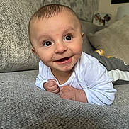 Khalil is registered to the contest to win money with this photo: baby, smiling, face, infant, couch, gray, white_clothing, hands, eyes, furniture, indoors, portrait, happy, toddler, skin, head, expression, background, wallpaper, cozy