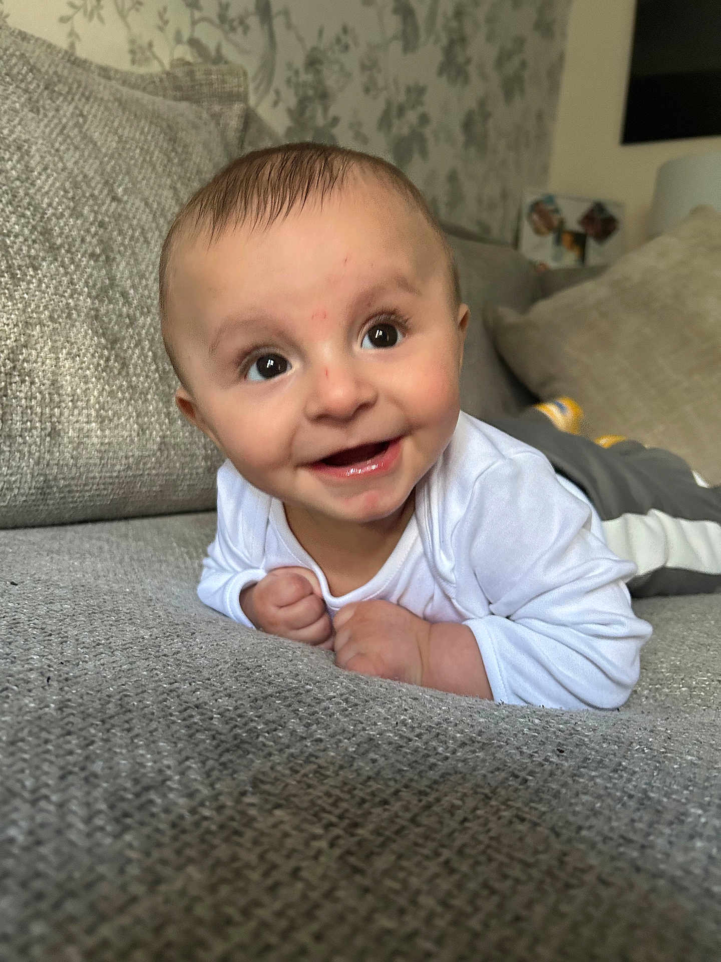 Khalil is registered to the contest to win money with this photo: baby, smiling, face, infant, couch, gray, white_clothing, hands, eyes, furniture, indoors, portrait, happy, toddler, skin, head, expression, background, wallpaper, cozy