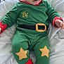 Khalil joined the competition — help win amazing prizes! baby, child, infant, green_outfit, elf_costume, red_accents, star_patches, footed_pajamas, toy, blanket, white_bedding, indoors, portrait, cute, holiday, festive, smiling, person, sitting, young