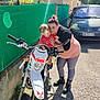 child, woman, motorbike, outdoor, sunlight, smile, fence, vehicle, car, road, person, casual_clothing, boots, short_hair, happy, embrace, urban, daytime, transportation, family