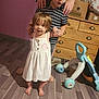 child, toddler, indoor, wooden_floor, dresser, toy, baby_dress, barefoot, person, supporting_hands, striped_shirt, flooring, home, family, playtime, young_children, childhood, cute, smiling, learning_to_walk