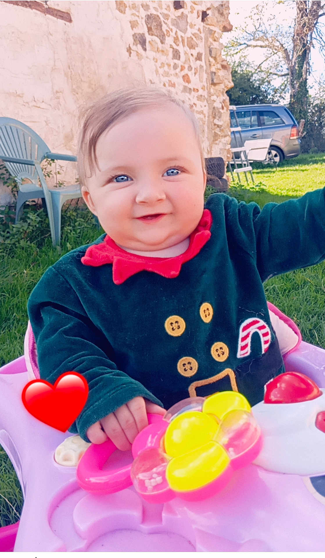 Lylyana participe au concours pour gagner de l'argent avec cette photo : baby, child, blue_eyes, smiling, outdoor, grass, walker, toy, pink, red_heart, festive_clothing, button_details, stone_wall, green_chair, sunny, daylight, happy, play, person, closeup