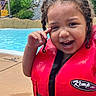 child, closeup, curly_hair, face, fun, greenery, happy, life_jacket, nature, outdoor, person, playful, pool, recreation, safety, smiling, summer, water, wet, winking