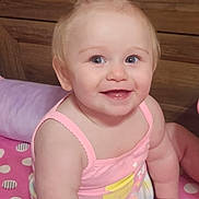 Bella is registered to the contest to win money with this photo: arm, baby, blanket, cheerful, child, cute, eyes, face, hand, happy, indoor, infant, pink_clothing, polka_dot, portrait, sitting, skin, smile, topknot, wooden_background
