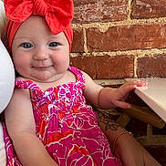 Amelia is registered to the contest to win money with this photo: baby, child, headband, red_bow, pink_dress, floral_pattern, smile, face, arm, bracelet, tattoo, brick_wall, indoor, person, cute, portrait, seated, hand, table, happy