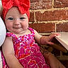 baby, child, headband, red_bow, pink_dress, floral_pattern, smile, face, arm, bracelet, tattoo, brick_wall, indoor, person, cute, portrait, seated, hand, table, happy