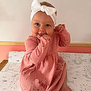 Penelope is registered to the contest to win money with this photo: baby, child, girl, smile, headband, bow, pink_dress, sitting, barefoot, indoor, changing_mat, patterned_mat, cute, happy, portrait, infant, playful, clothing, face, hand
