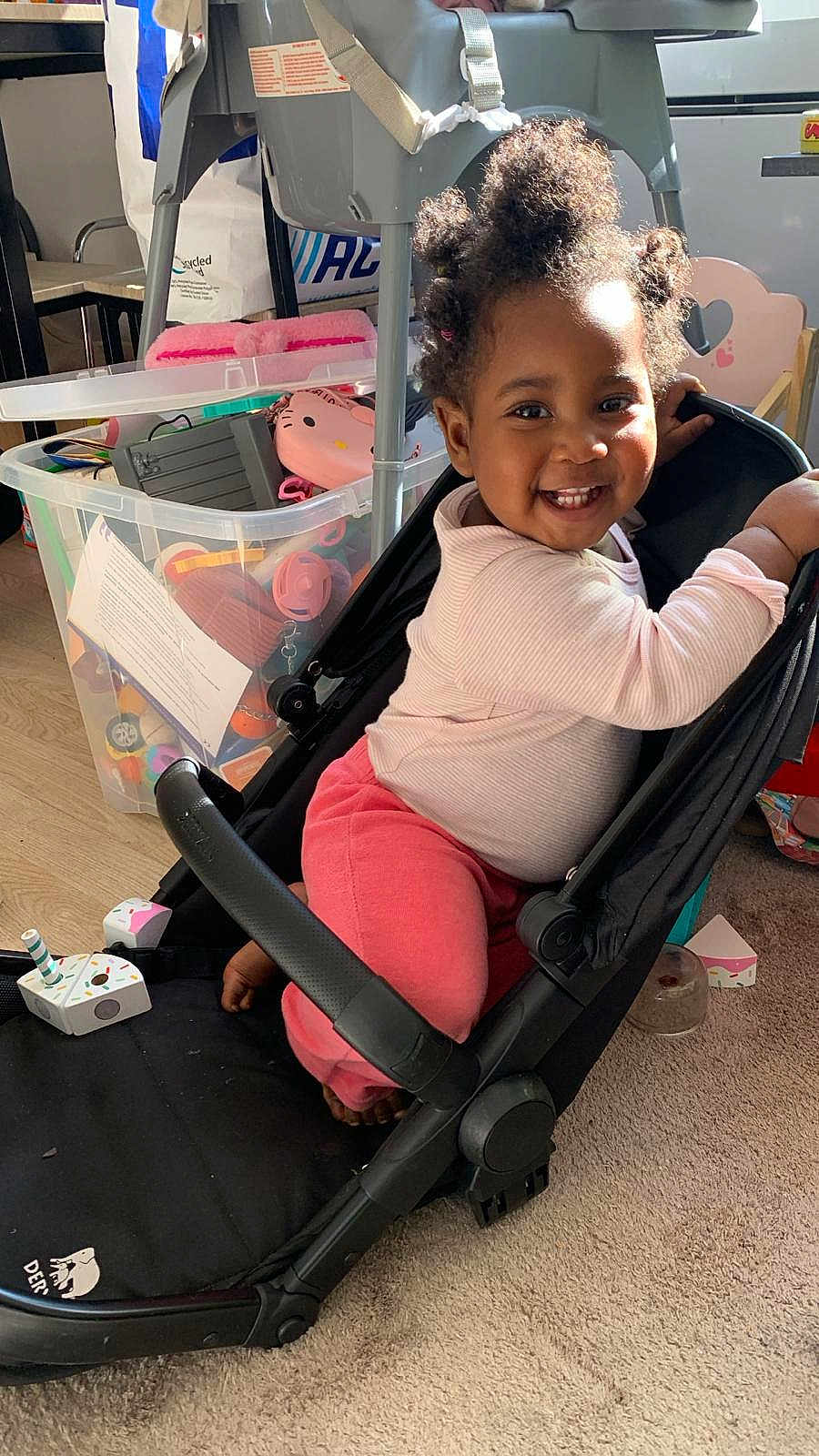 Ariana participe au concours pour gagner de l'argent avec cette photo : child, toddler, smile, happy, stroller, toys, toy_bin, indoor, living_room, sunlight, curly_hair, pink_pants, white_shirt, carpet, floor, seat, playtime, highchair, container, portrait