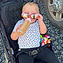 toddler, stroller, sunglasses, bottle, toy, gravel, wheel, black_clothing, white_shirt, red_shoes, child, outdoor, person, baby, seatbelt, headwear, playful, casual, reflection, cute