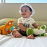 Alonzo joined the competition — help win amazing prizes! baby, child, sitting, smiling, sunhat, bib, onesie, barefoot, stuffed_animal, dinosaur_toy, orange_toy, green_toy, bed, blanket, bedroom, window, indoor, portrait, happy, cozy