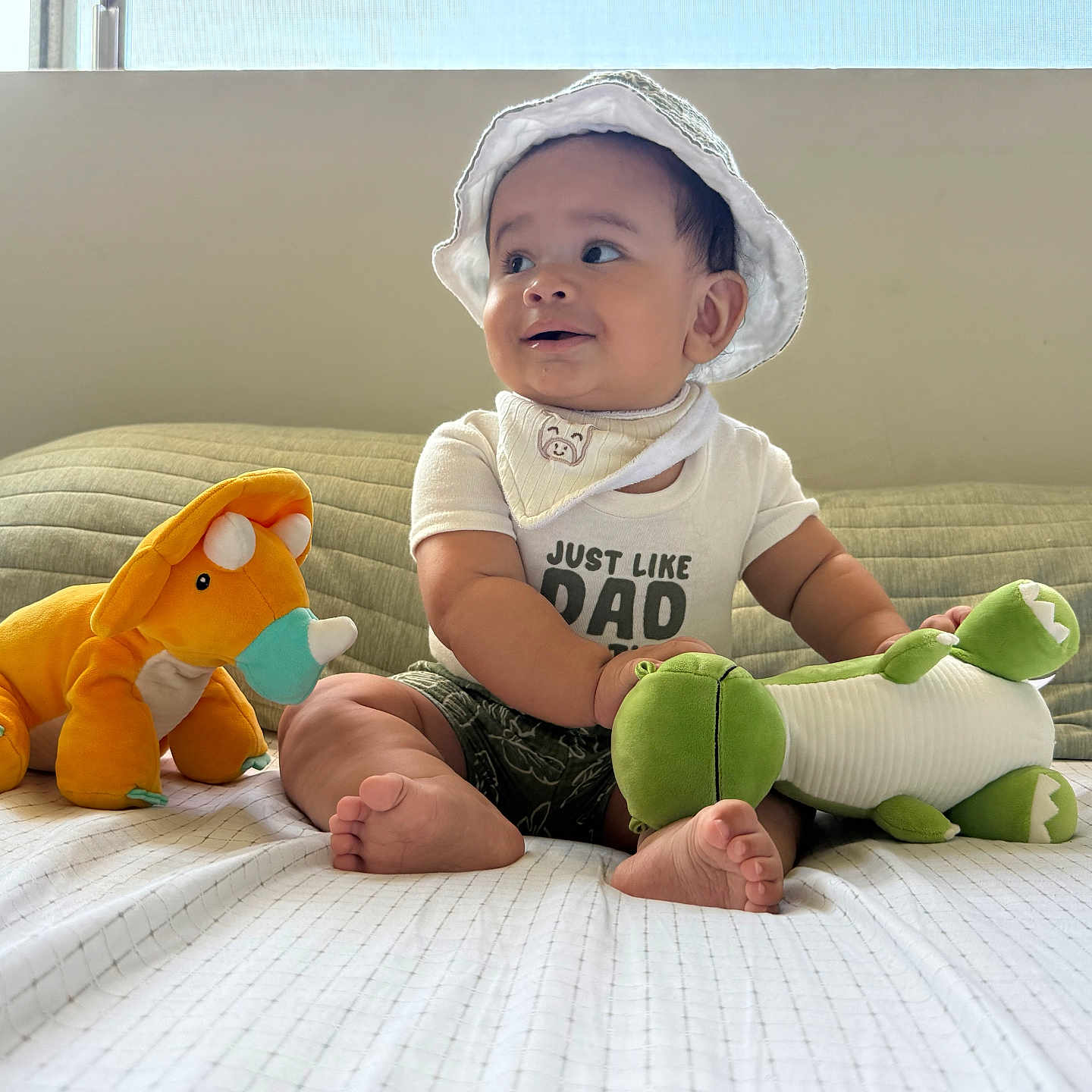 Alonzo joined the competition — help win amazing prizes! baby, child, sitting, smiling, sunhat, bib, onesie, barefoot, stuffed_animal, dinosaur_toy, orange_toy, green_toy, bed, blanket, bedroom, window, indoor, portrait, happy, cozy