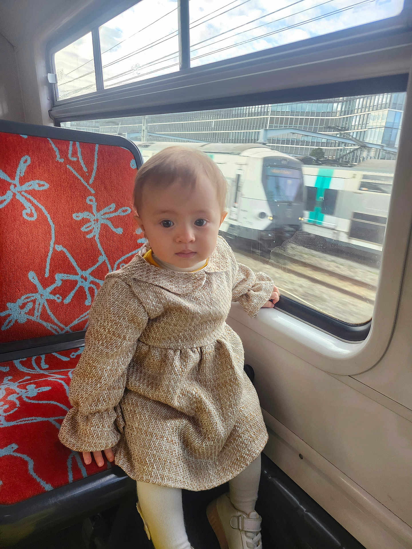 Sara participe au concours pour gagner de l'argent avec cette photo : toddler, child, train, window, seat, dress, beige, red, pattern, transport, public_transport, railway, travel, curious, indoors, person, footwear, white_shoes, looking, daylight