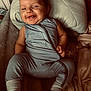 baby, smiling, blue_onesie, pillow, blanket, remote_control, infant, happy, lying_down, cozy, soft_texture, indoor, cute, child, portrait, socks, relaxed, candid, warm_lighting, face