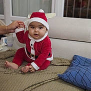 Lyna participe au concours pour gagner de l'argent avec cette photo : baby, child, santa_outfit, red_clothing, hat, indoor, couch, pillow, hand, holding, person, infant, smiling, face, cute, holiday, festive, home, blanket, seated