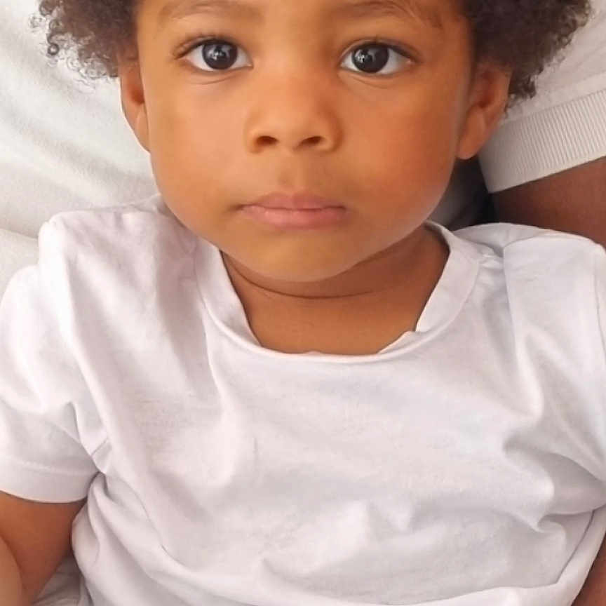 Moïse a rejoint le concours — aidez-le/la à gagner de superbes lots ! adult_arm, afro_hair, candid, casual_clothing, child, closeup, comfort, curious, expression, eyes, face, human, indoor, person, portrait, seated, skin, toddler, white_tshirt, young_child