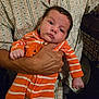 Nicolas is registered to the contest to win money with this photo: baby, infant, person, hand, clothing, striped_onesie, orange_color, pumpkin_design, adult, holding, patterned_shirt, face, expression, indoors, portrait, cute, newborn, skin, head, human