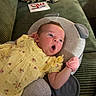 baby, yellow_dress, pink_mitten, cushion, green_couch, leopard_print_pillow, bag, book, face, infant, expression, plush, person, child, indoor, comfort, resting, closeup, soft_fabric, home