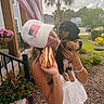 Tucker is registered to the contest to win money with this photo: woman, puppy, dog, hot_dog, cap, american_flag, porch, flowers, plants, greenery, outdoor, daytime, casual_clothing, smiling, holding, pet, nature, residential, house, summer