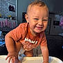 toddler, child, smiling, orange_shirt, high_chair, indoor, home, balloons, family_photo, playful, cute, cheeky, baby, furniture, hands, feet, diaper, person, portrait, happy
