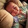 baby, infant, sleeping, pacifier, pillow, blanket, onesie, smile, face, close_up, cozy, bedtime, bedding, pacifier_clip, bead_teether, chubby_cheeks, snuggling, indoor, portrait, calm