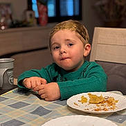 Gabin participe au concours pour gagner de l'argent avec cette photo : child, toddler, boy, green_sweater, headband, paper_plate, crumbs, food, tablecloth, dining_table, cup, chair, window, blurred_background, portrait, candid, meal_time, face, hands, snack