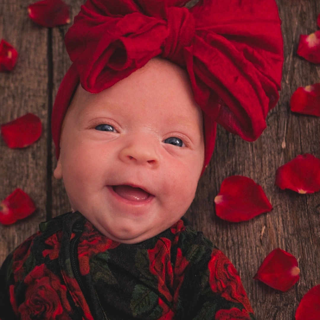 Saoirse joined the competition — help win amazing prizes! baby, bow, cheeks, closeup, cute, eyes, fabric, floral_outfit, happy, headwear, headwrap, infant, newborn, portrait, red, rose_petals, rustic, smiling, texture, wooden_floor