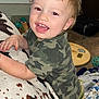 baby, blanket, blonde_hair, camouflage_clothing, carpet, child, couch, cute, expression, face, furniture, happy, home, indoor, pattern, person, playful, smiling, toddler, toy